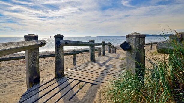 Eine Holzpromenade führt zu einem Sandstrand mit freiem Blick auf das Meer und das entfernte Land.