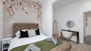 Ein Schlafzimmer mit einer gepolsterten Kopfteil, einem Bett mit weißer Bettwäsche, einer grünen Tagesdecke und gerollten Handtüchern. Ein weißer Stuhl, eine Holzleiter und ein runder Spiegel sind an der Wand angebracht.