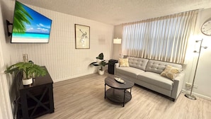 A modern living room with a sofa, coffee table, and a TV displaying a beach scene. - Relaxing Kalakauan 1-Bedroom | Recently Redecorated! (Honolulu)