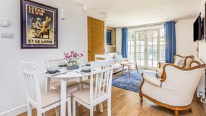 A dining area with a white table set for four, a white sofa, and a vintage armchair. A framed poster is on the wall, and a blue curtain is partially drawn. - Charming Cottage with Garden - Pass the Keys (Chichester)