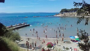 Een strand met mensen die zwemmen en zonnebaden, een pier met boten en een rotsachtige kustlijn.