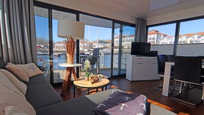 Habitación de hotel moderna con un ventanal amplio que ofrece vista al puerto deportivo, un sofá, una mesita redonda y un escritorio con un televisor.
