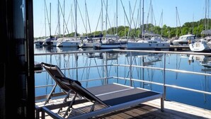 A marina with multiple sailboats docked, a lounge chair on a wooden deck, and a clear view of the water.