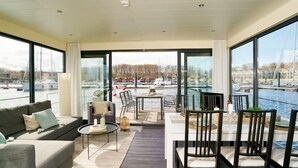 A modern living room with a sofa, coffee table, and dining area with chairs. Large windows offer views of a marina with boats. - Hausboot in Boltenhagen (Boltenhagen)