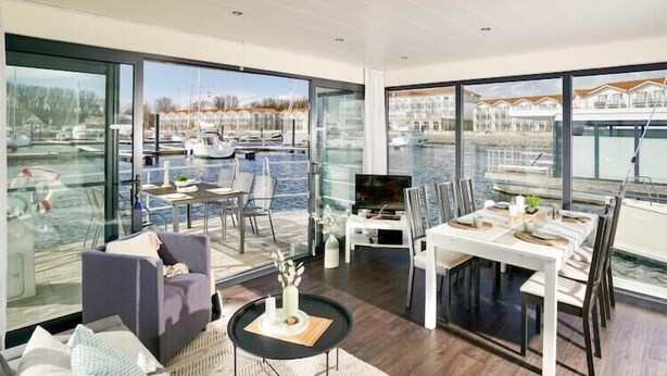 A spacious living area with a large window overlooking a marina, a dining table set for six, and a comfortable seating area with a sofa and chairs. - Hausboot in Boltenhagen (Boltenhagen)