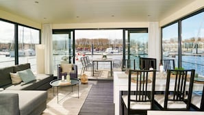 Ein modernes Wohnzimmer mit Sofa, Couchtisch und Essbereich mit Stühlen. Große Fenster bieten Blick auf eine Marina mit Booten.