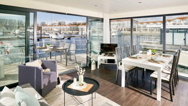 Ein modernes Wohnzimmer mit einer großen Glaswand mit Blick auf eine Marina, einem Essbereich mit weißer Möbel und einer gemütlichen Sitzecke mit Sofa und Couchtisch.
