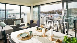 Ein modernes Zimmer mit großem Fenster mit Blick auf eine Marina, inklusive Sofa, Esstisch gedeckt mit Tellern, Gläsern und Kerzen.