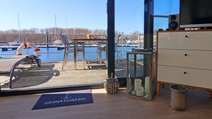 A room with a view of a marina, a wooden deck, a desk, a chair, a small table with candles, and a blue mat with text.