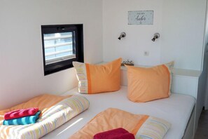Une chambre avec deux lits, chacun agrémenté de draps à rayures et d’oreillers orange. On y trouve une fenêtre avec des stores, une petite étagère avec des livres et une horloge murale.