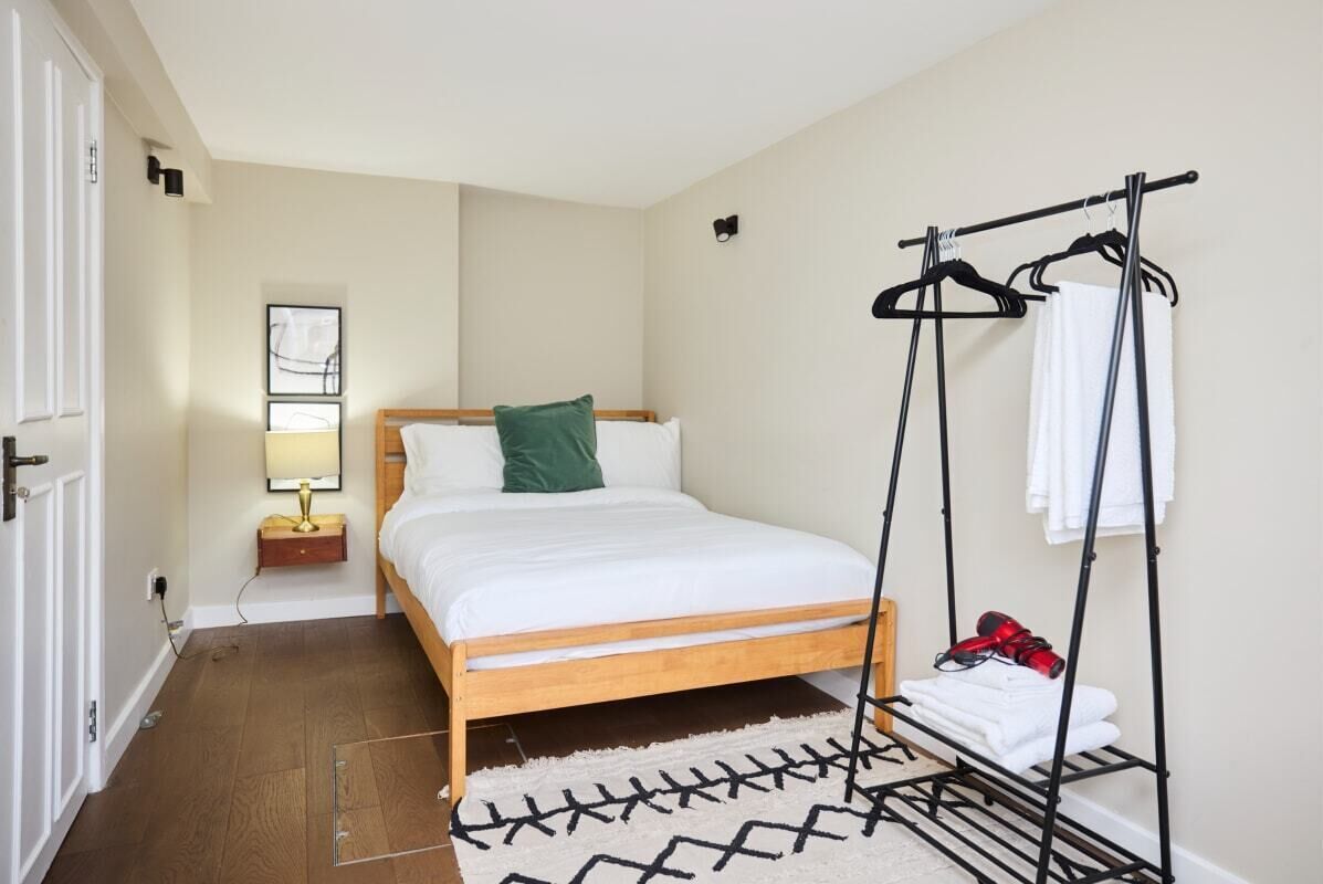A bedroom with a wooden bed, a bedside table with a lamp, a wall-mounted drying rack, and a door.
