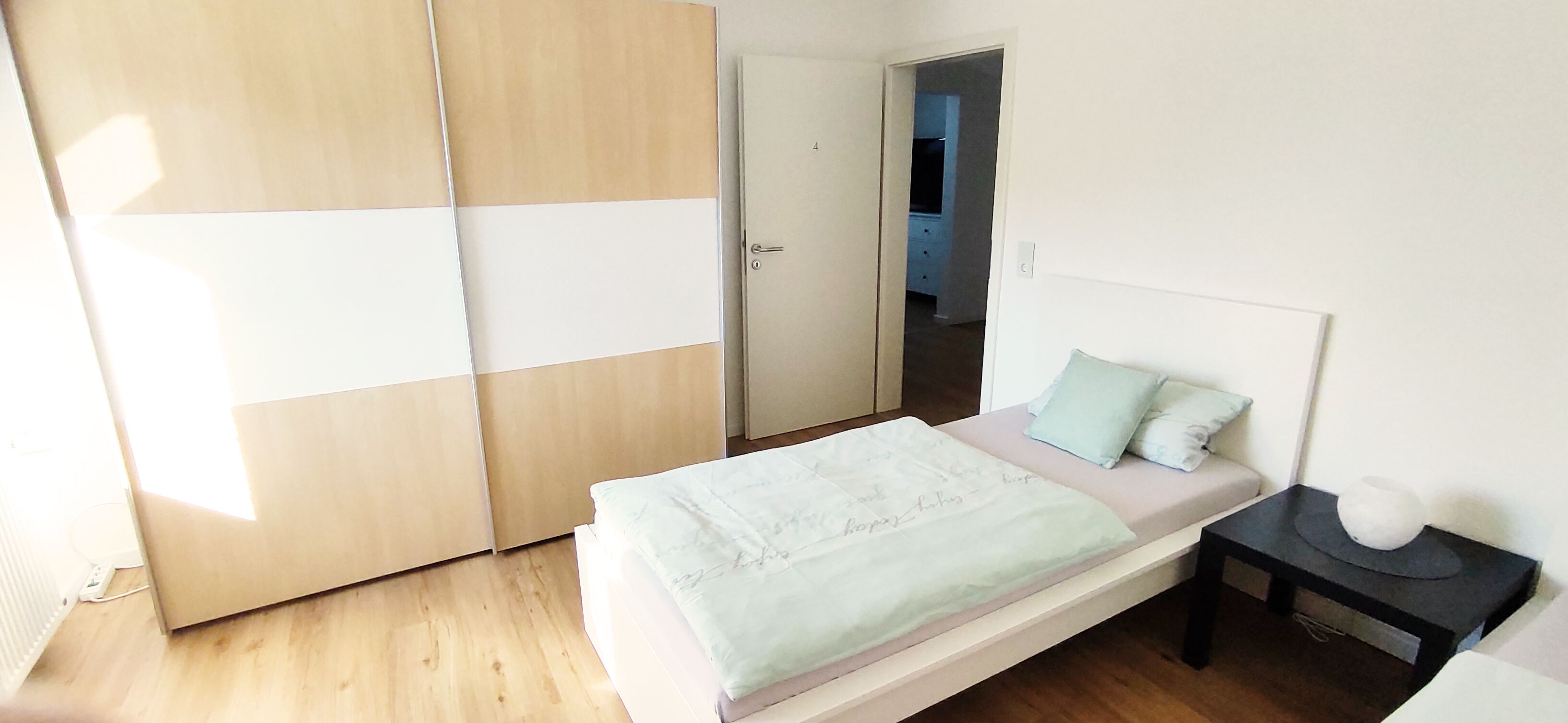 A bedroom with a bed, a nightstand, a wardrobe, and a door leading to another room.