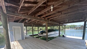 Une terrasse en bois couverte, avec un bateau amarré en dessous, une porte et une vue sur une étendue d’eau, avec des bateaux et de la végétation en arrière-plan.