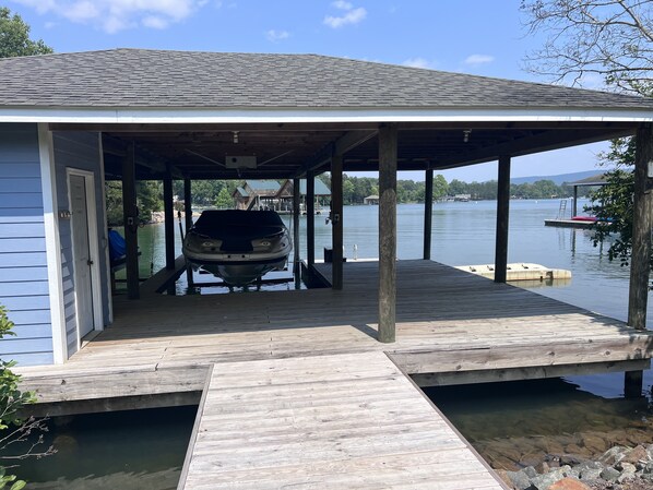 A covered boat dock with a boat inside, a wooden deck, and a view of a lake with buildings in the background.