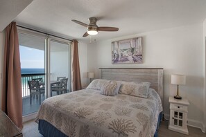 Ein Schlafzimmer mit Bett, Deckenventilator, einem Gemälde, einem Nachttisch und Meerblick durch eine Schiebetür.