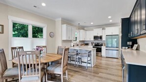 A modern kitchen with white cabinets, a central island, and a dining area with wooden chairs and a table. - The Sand-Bar (Carolina Beach)