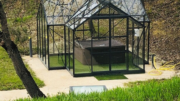 A glass greenhouse structure with a metal frame, surrounded by grass and trees.