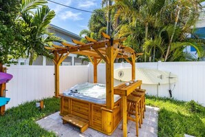 Relax in a hot tub under a shaded pergola, surrounded by lush greenery and a palm tree.
