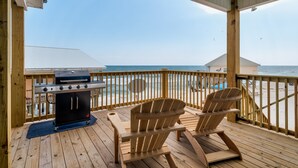 A wooden deck with a grill and two Adirondack chairs overlooking the beach.