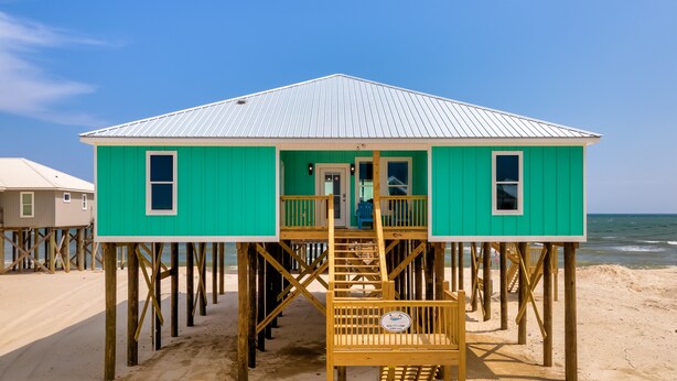 A turquoise beach house with a metal roof, wooden deck, and a staircase leading to the entrance.