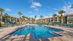 A swimming pool surrounded by palm trees and lounge chairs, with a hotel building in the background. - Ocean Villas 6 2 Bedroom Condo by RedAwning (St. Augustine)