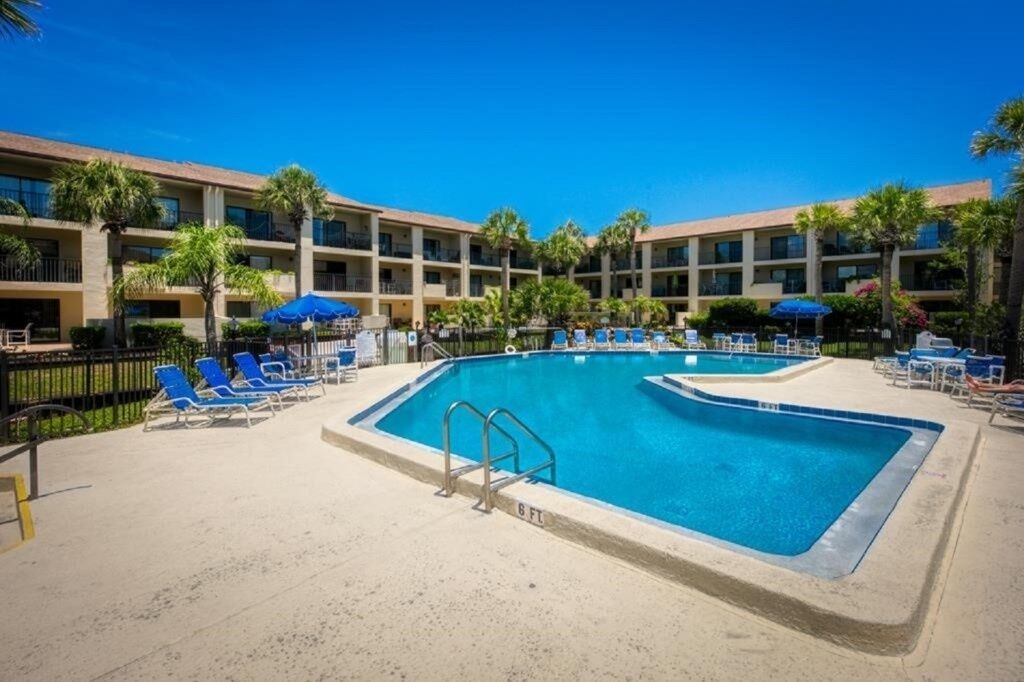 A hotel pool area with multiple lounge chairs and umbrellas, surrounded by a multi-story building with balconies.