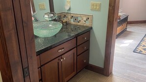 A bathroom with a glass bowl sink, wooden cabinets, a tiled backsplash, and a pebbled floor. - This casita is located in the Estrella neighborhood and boasts amazing View’s.  (Goodyear)