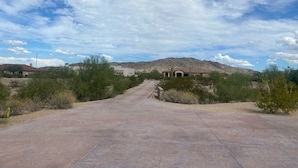A paved road leading to a building with a flat roof and a small porch, surrounded by sparse vegetation and a hilly landscape under a partly cloudy sky. - This casita is located in the Estrella neighborhood and boasts amazing View’s.  (Goodyear)