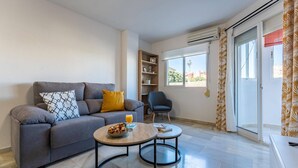 A modern living room with a grey sofa, a small round table with a bowl of snacks, a chair, and a bookshelf.