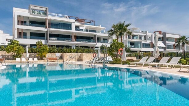 A modern hotel with a large swimming pool, sun loungers, and palm trees.