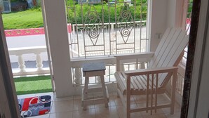 A porch with a white chair and a small table, a metal gate, and a view of a green lawn and a red and white fence. - Lovely 1 bedroom, 2 Guest +, holiday rentals in St Thomas Jamaica  (Port Morant)