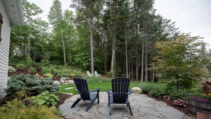 Un patio de piedra con dos sillas Adirondack mirando hacia un jardín exuberante y árboles altos.