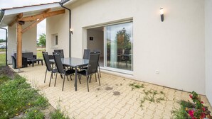 En udendørs terrasse med et bord, stole, en pergola og en glasdør ind til et overdækket område.