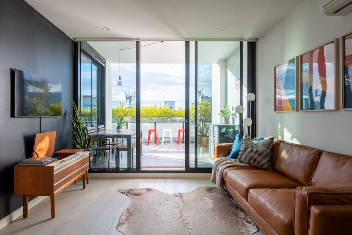 A modern living room with a leather sofa, a wooden desk, and a glass sliding door leading to an outdoor balcony.