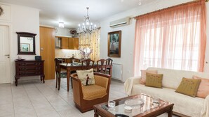 A living room with a sofa, chairs, a coffee table, a dining table with chairs, a chandelier, and a large window with sheer curtains. - Deluxe 2 Bedroom apt in Petroupoli (Petroupoli)
