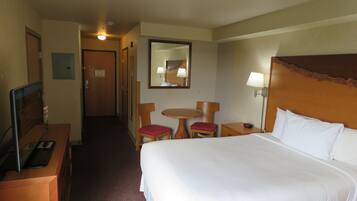A hotel room with a large bed, a desk with a computer, a chair, a small table, and a lamp.