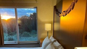 Habitación de hotel con cama, mesita de noche, lámpara y vistas al exterior.