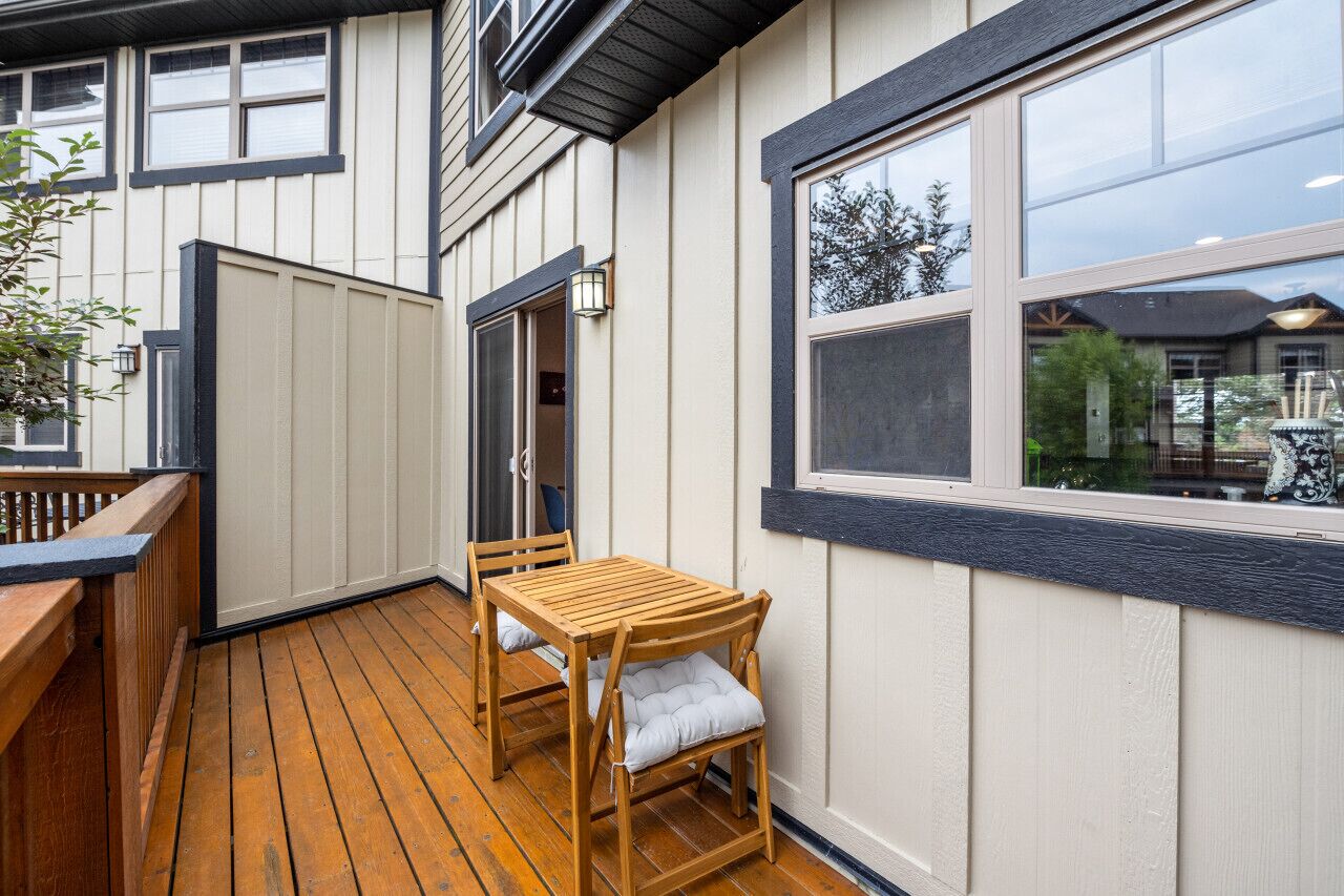 Um deck de madeira com mesa e cadeiras, uma porta que dá para o interior e uma janela grande com vista para o exterior.