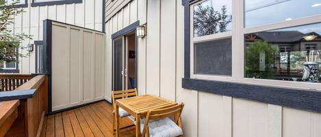 Um deck de madeira com mesa e cadeiras, uma porta que dá para o interior e uma janela grande com vista para o exterior.