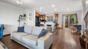 Una sala de estar moderna con un sofá, una zona de comedor con taburetes, una cocina con armarios de madera y un ventanal grande.