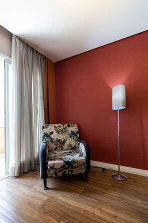 A room with a red wall, a floral-patterned armchair, a floor lamp, and a window with curtains.