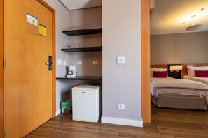 A compact hotel room with a wooden door, a small refrigerator, a kitchenette with shelves, a bed with red pillows, and a bedside table. - Elegance Suites Stella Vega (São Paulo)