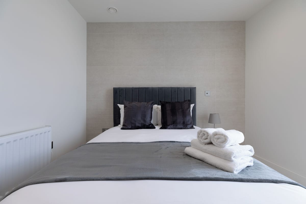 A modern hotel room with a large bed, grey bedding, a dark headboard, and a stack of white towels.