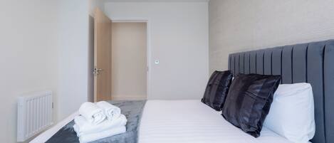A modern hotel room with a neatly made bed, a radiator, and a door leading to another room.