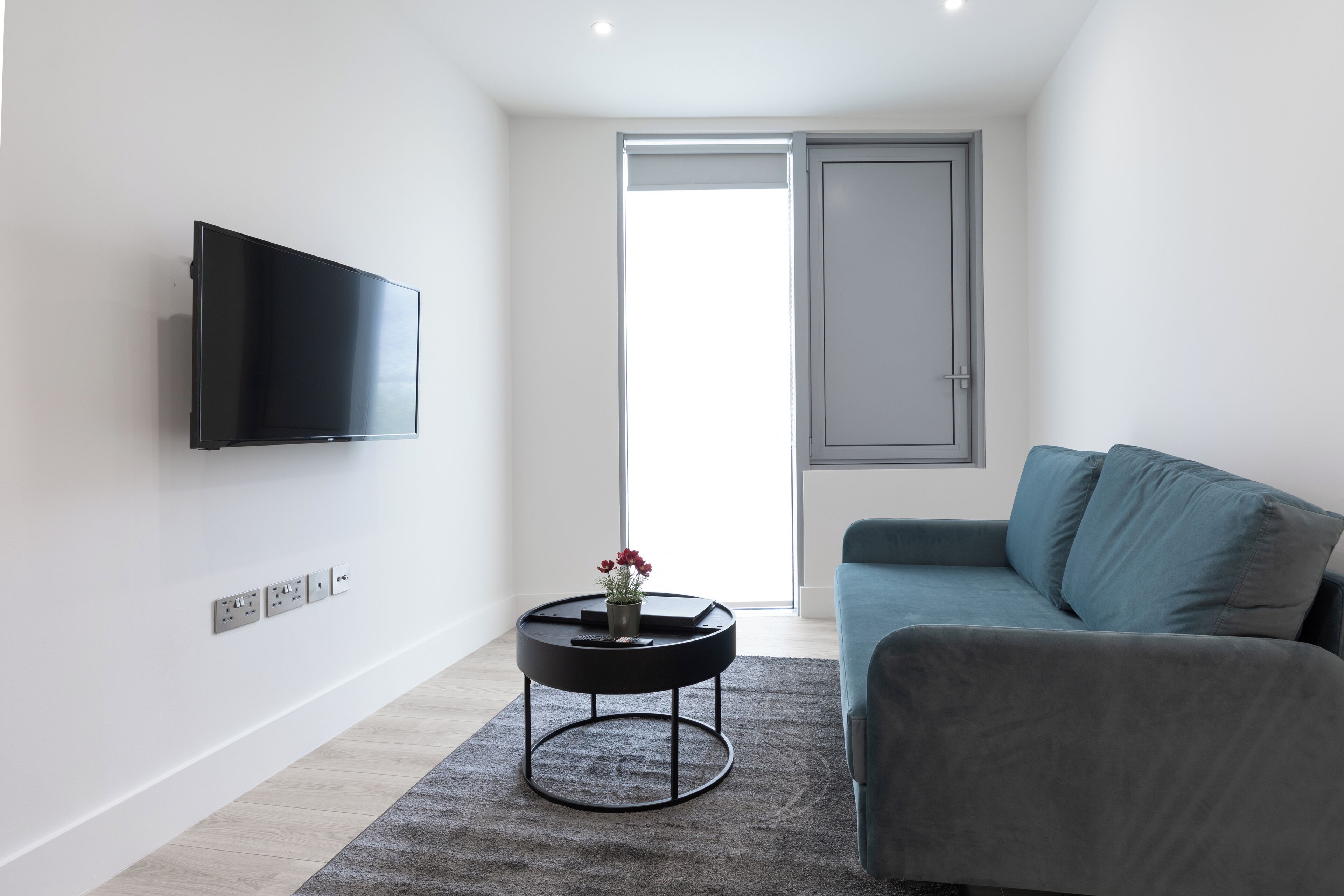 A modern living room with a flat-screen TV mounted on the wall, a round black coffee table with a vase of flowers, a grey sofa, and a door leading to a bright room.