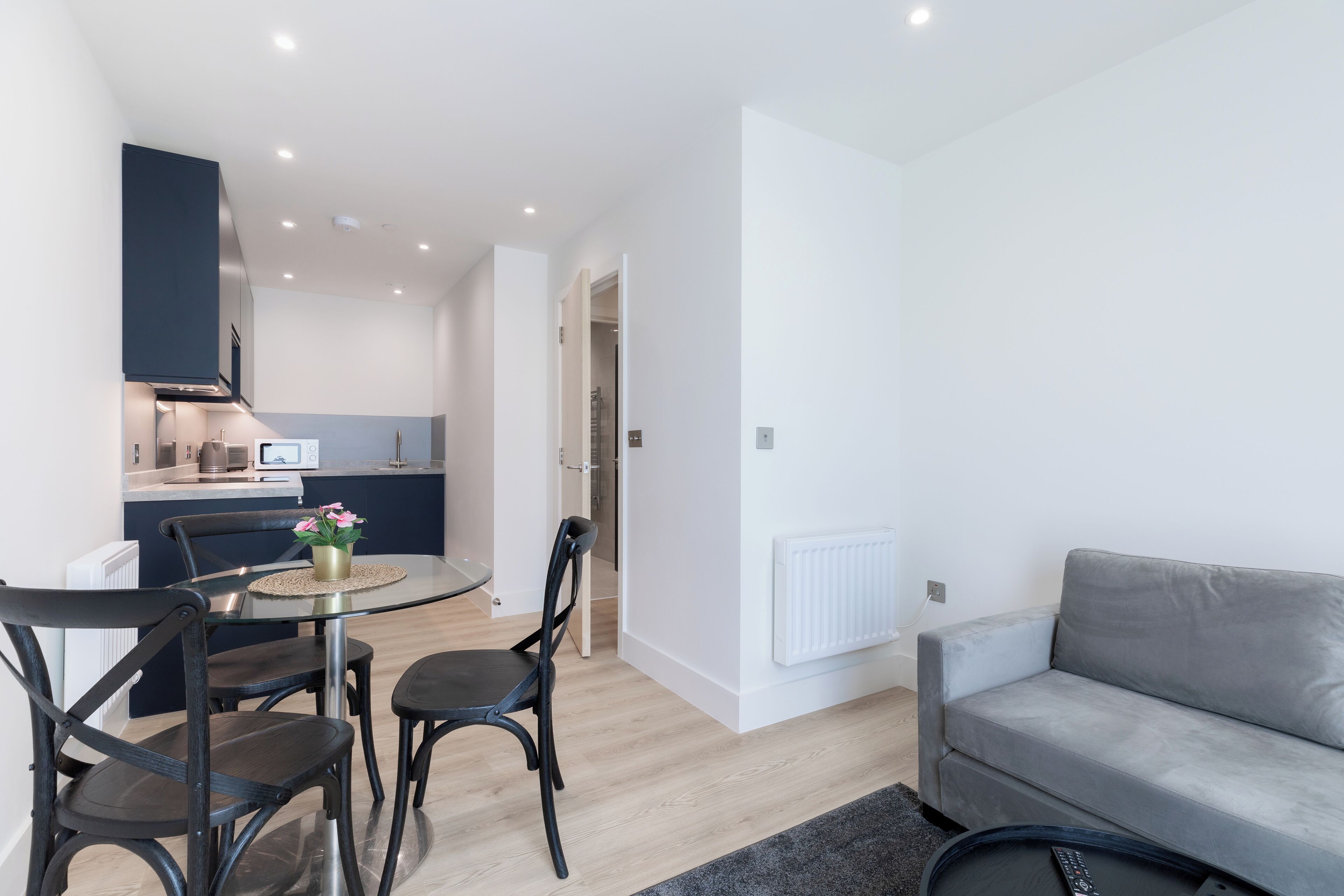 A modern living room with a grey sofa, a dining table with four chairs, and a kitchen in the background.