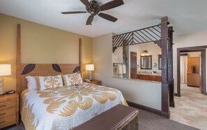 Ein Hotelzimmer mit einem großen Bett, einem Deckenventilator, einem hölzernen Kopfteil, einem Nachttisch mit Lampe, einer Bank und einem durch eine Glastür einsehbaren Badezimmer.