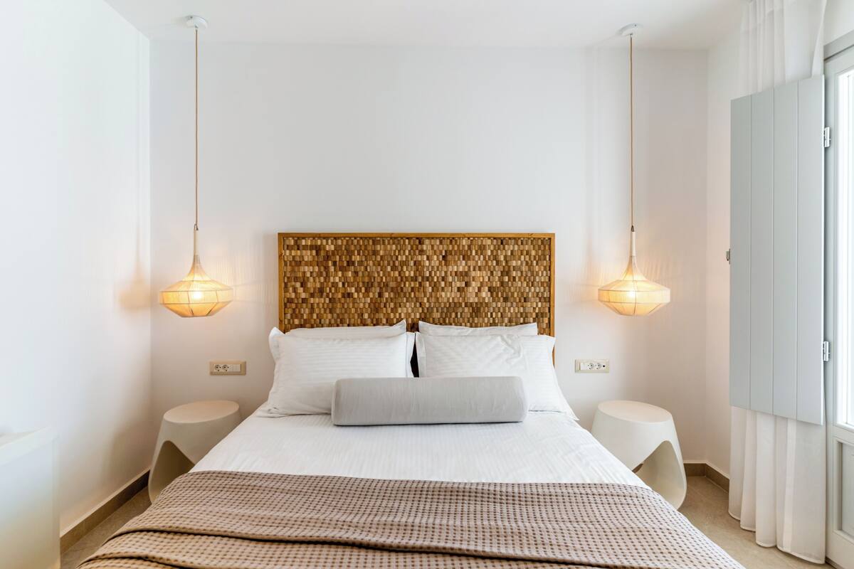 Una habitación de hotel moderna con una cama grande, dos mesitas de noche, un cabecero tejido e iluminación colgante.