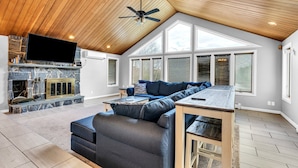 A spacious living room with a stone fireplace, a large flat-screen TV, a dark blue sectional sofa, and a wooden ceiling fan.
