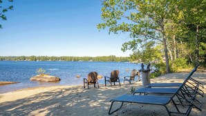 A lakeside beach with chairs and lounge chairs, a sandy shore, and a clear view of the water.